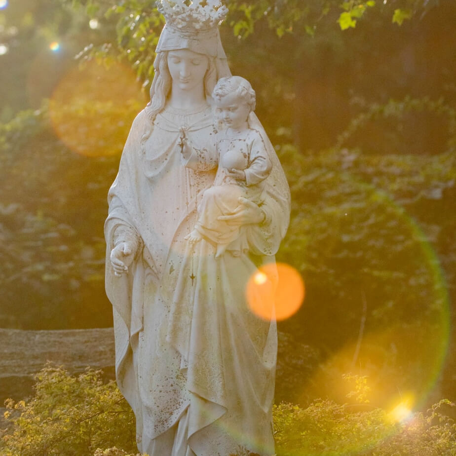 A sunlit statue of Mary cradling a child, radiating warmth and serenity in a peaceful outdoor setting.